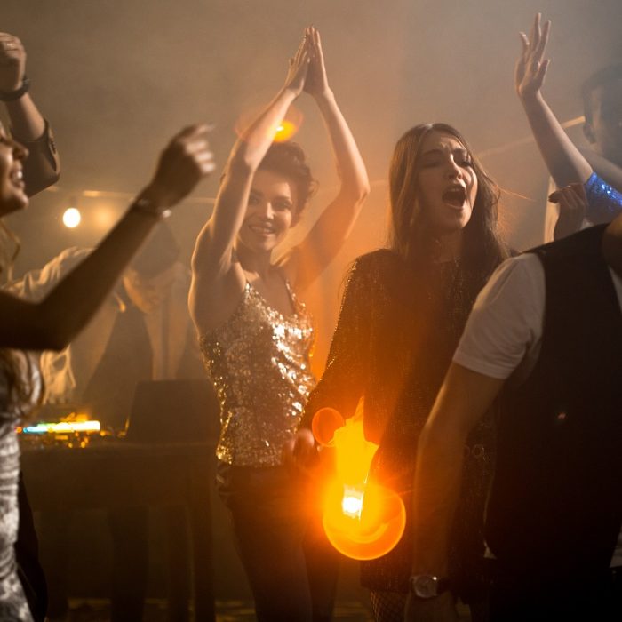 Crowd of trendy young people having fun in raving nightclub party, beautiful girls dancing on dim smoky dance floor